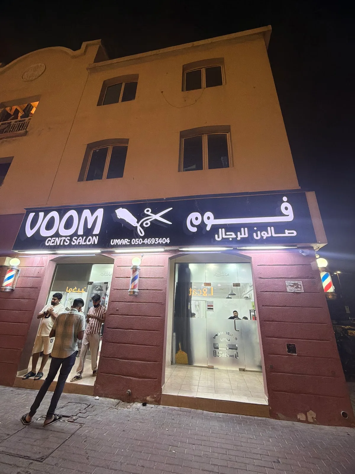 VOOM Gents Salon lit up at night, with barber poles flanking the entrance in Warsan First