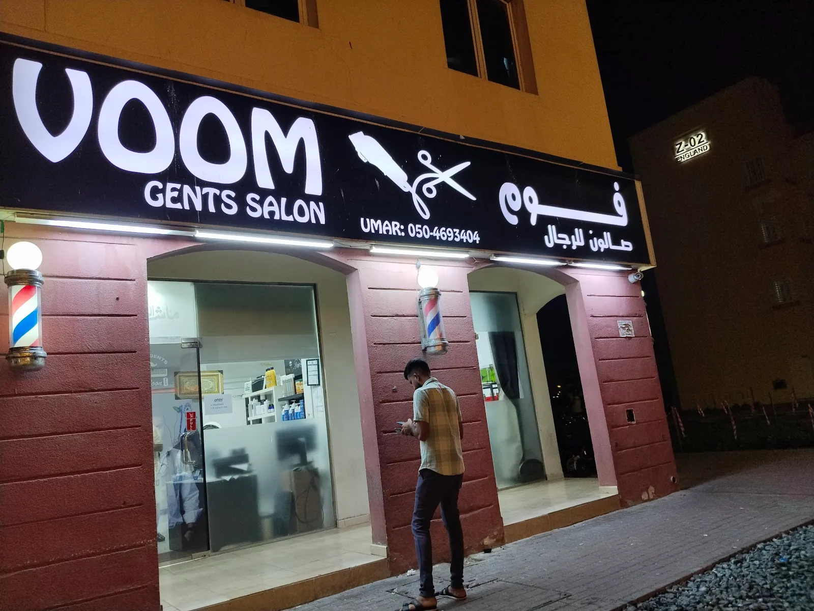 VOOM Gents Salon storefront at night with its illuminated wordmark sign and barber poles, England Cluster, Warsan First, Dubai
