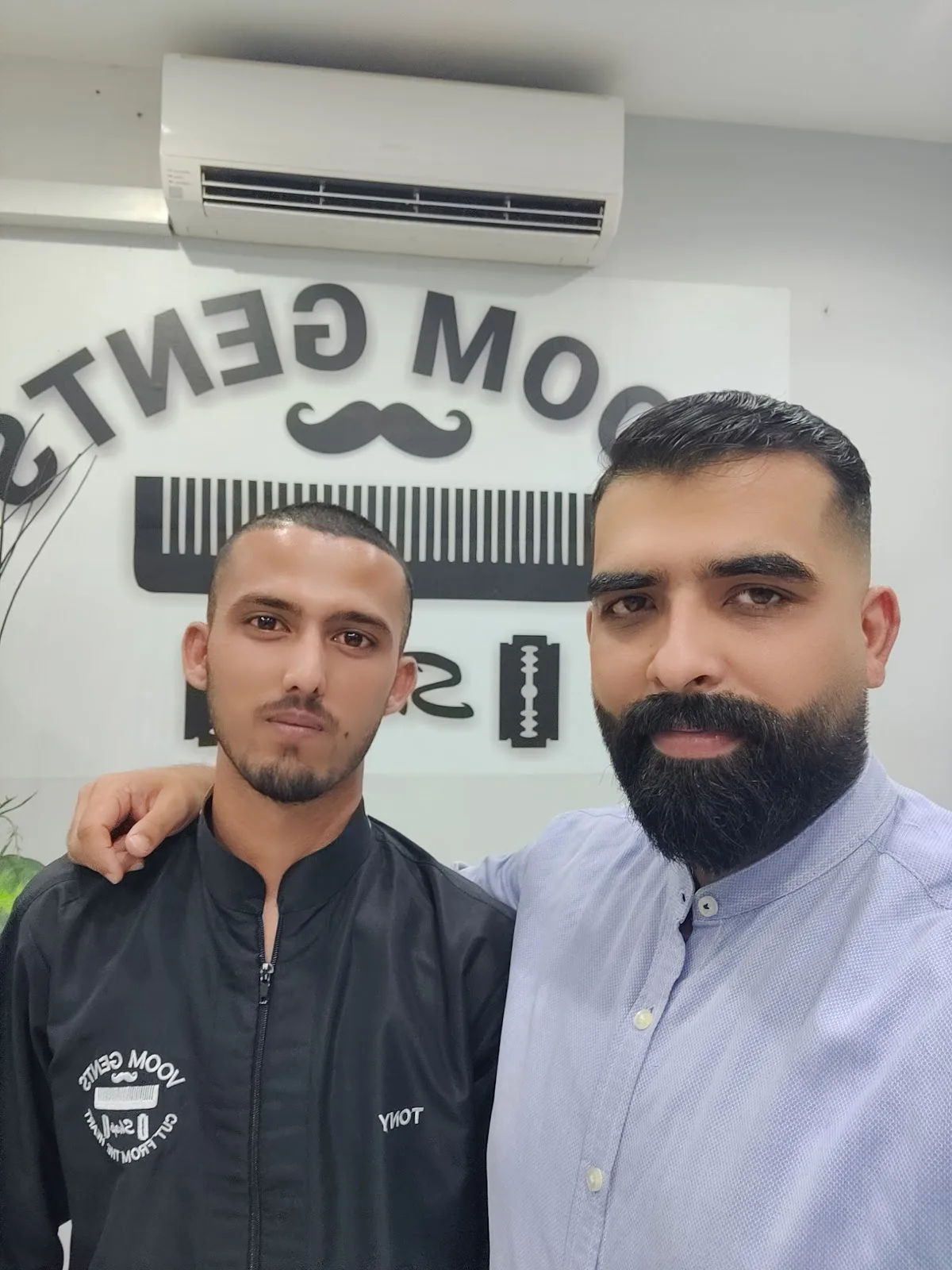 VOOM Gents Salon barber with a happy customer in front of the salon's comb-and-razor wall emblem